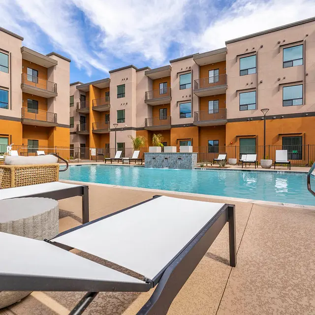 Modern apartment complex with a pool, lounge chairs, and a sunny sky in the background. Outdoor space is inviting and well-maintained.