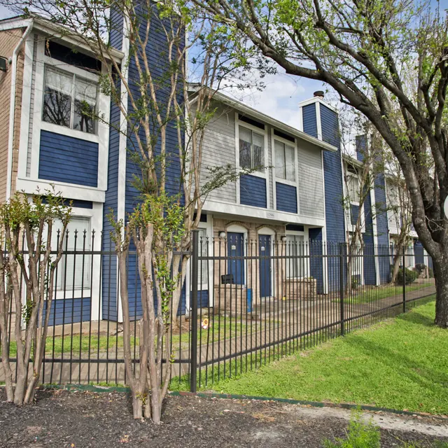 A modern two-story apartment building with blue and beige siding, surrounded by a black metal fence and green lawn. There are trees nearby, and a street lamp in the foreground.