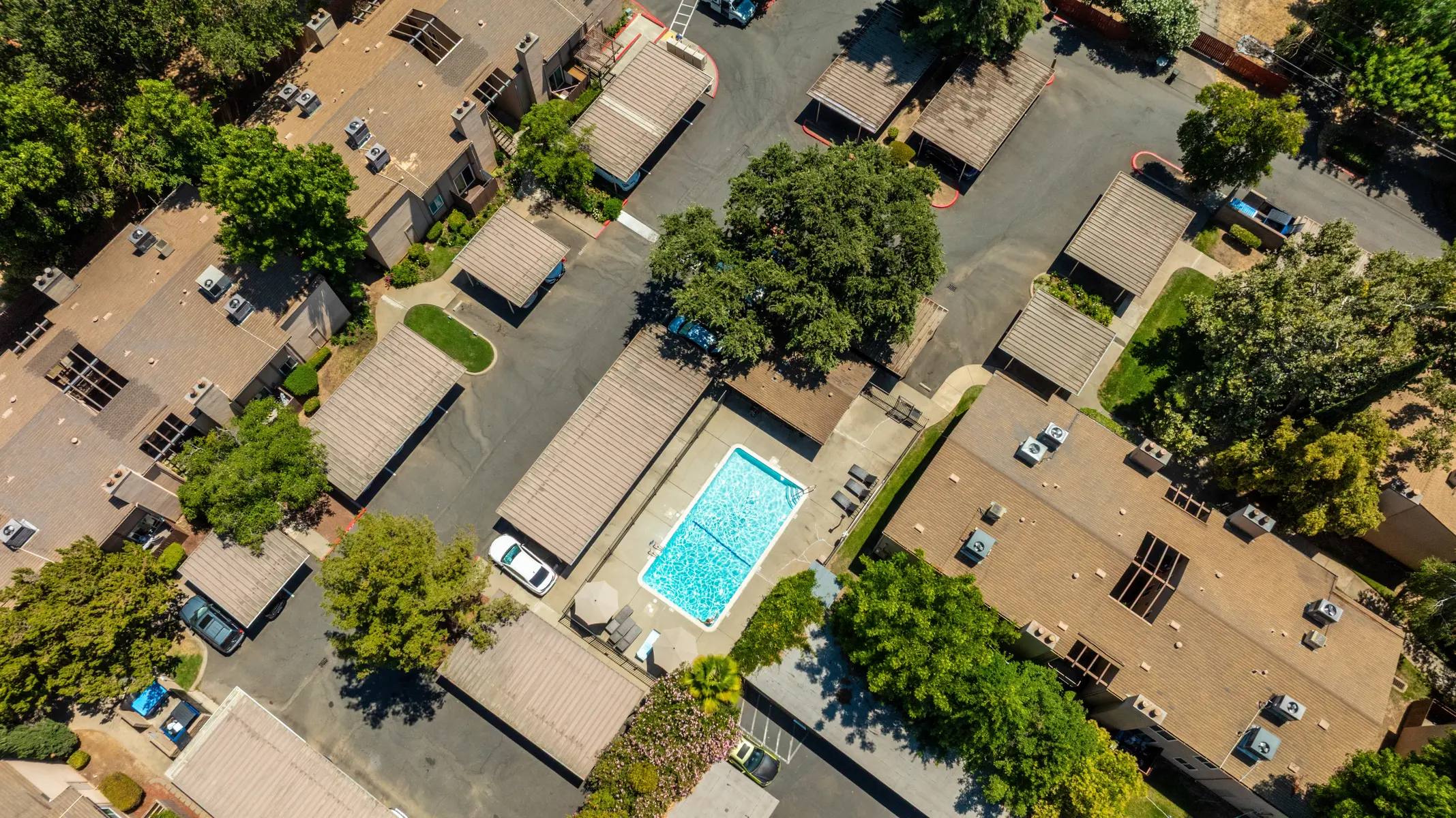 Aerial view of an apartment complex featuring a swimming pool surrounded by trees and parked cars.