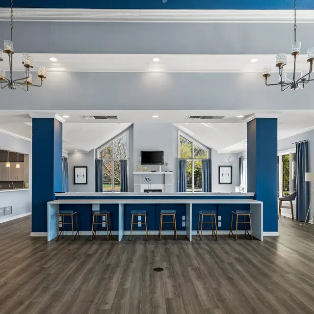 A spacious lounge area featuring a blue accent wall, a long white counter with bar stools, and large windows allowing natural light to enter. The floor is made of wood, and there are several chandeliers hanging from the ceiling.