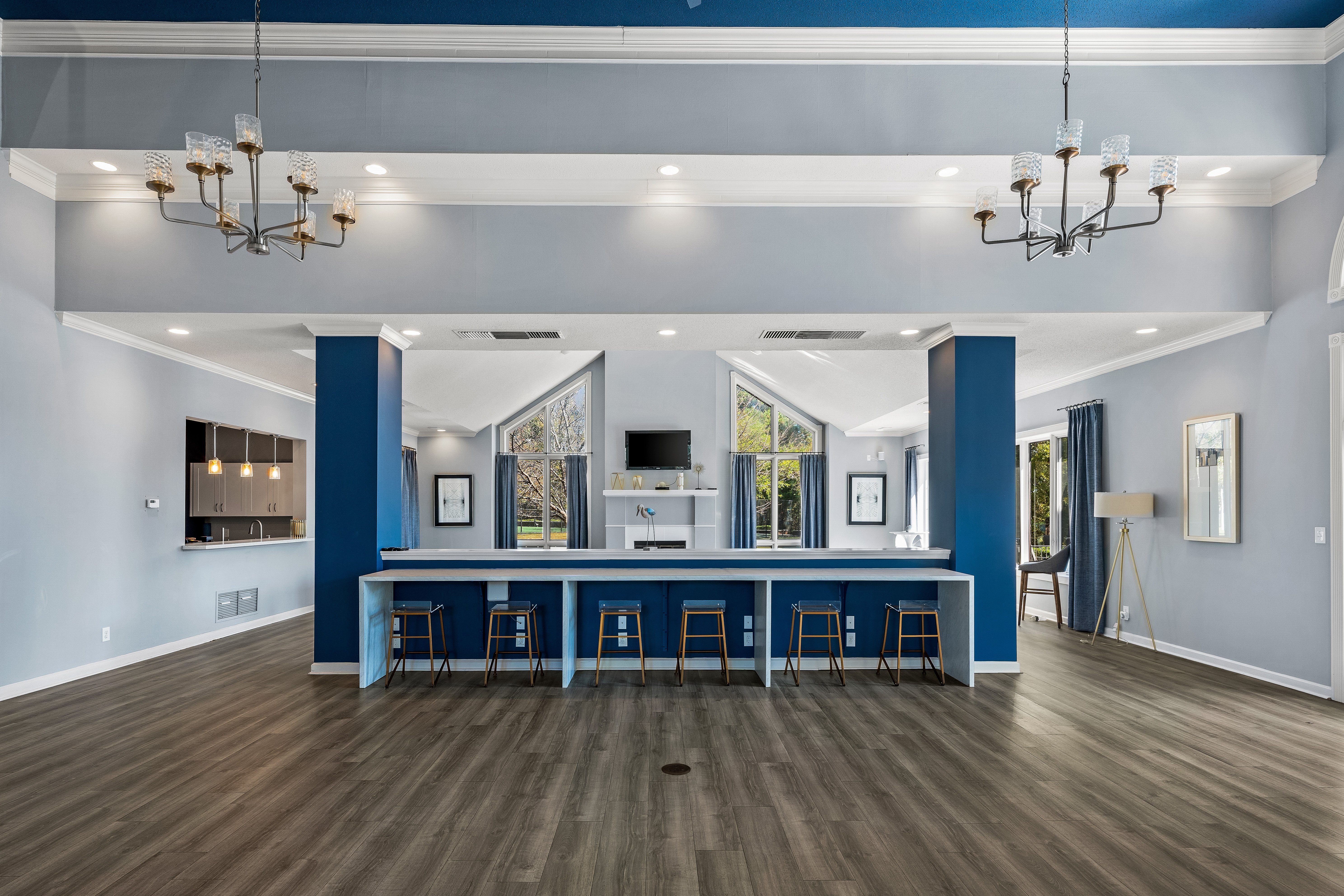 Modern Lounge Area A spacious lounge area featuring a blue accent wall, a long white counter with bar stools, and large windows allowing natural light to enter. The floor is made of wood, and there are several chandeliers hanging from the ceiling.