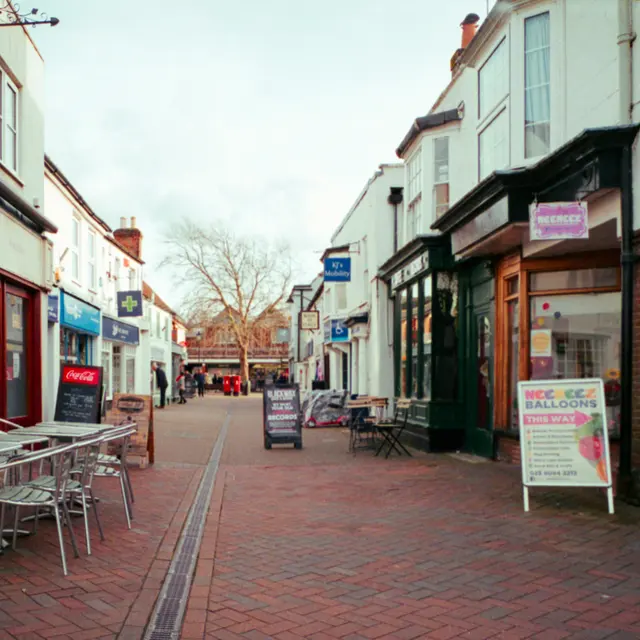 A quaint town street featuring shops on either side, with outdoor seating visible, and people walking in the distance.