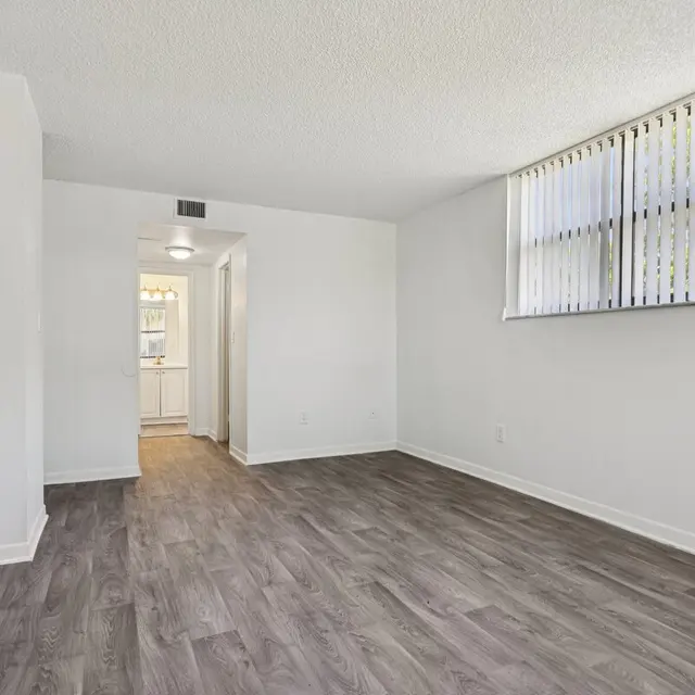 An empty apartment living room with light-colored walls and dark laminate flooring. Natural light comes from a window with vertical blinds. A doorway leads to another room.