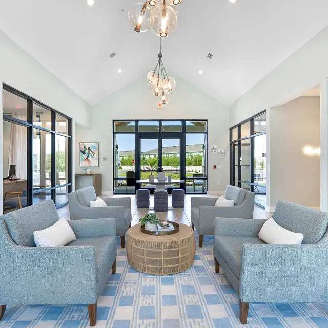 Stylish Lobby Interior A modern lobby interior featuring four upholstered chairs with cushions arranged around a round coffee table on a patterned area rug. Large windows allow natural light to fill the space, showing a pleasant outdoor view. The walls are painted in a light color and decorative light fixtures hang from the high ceiling.