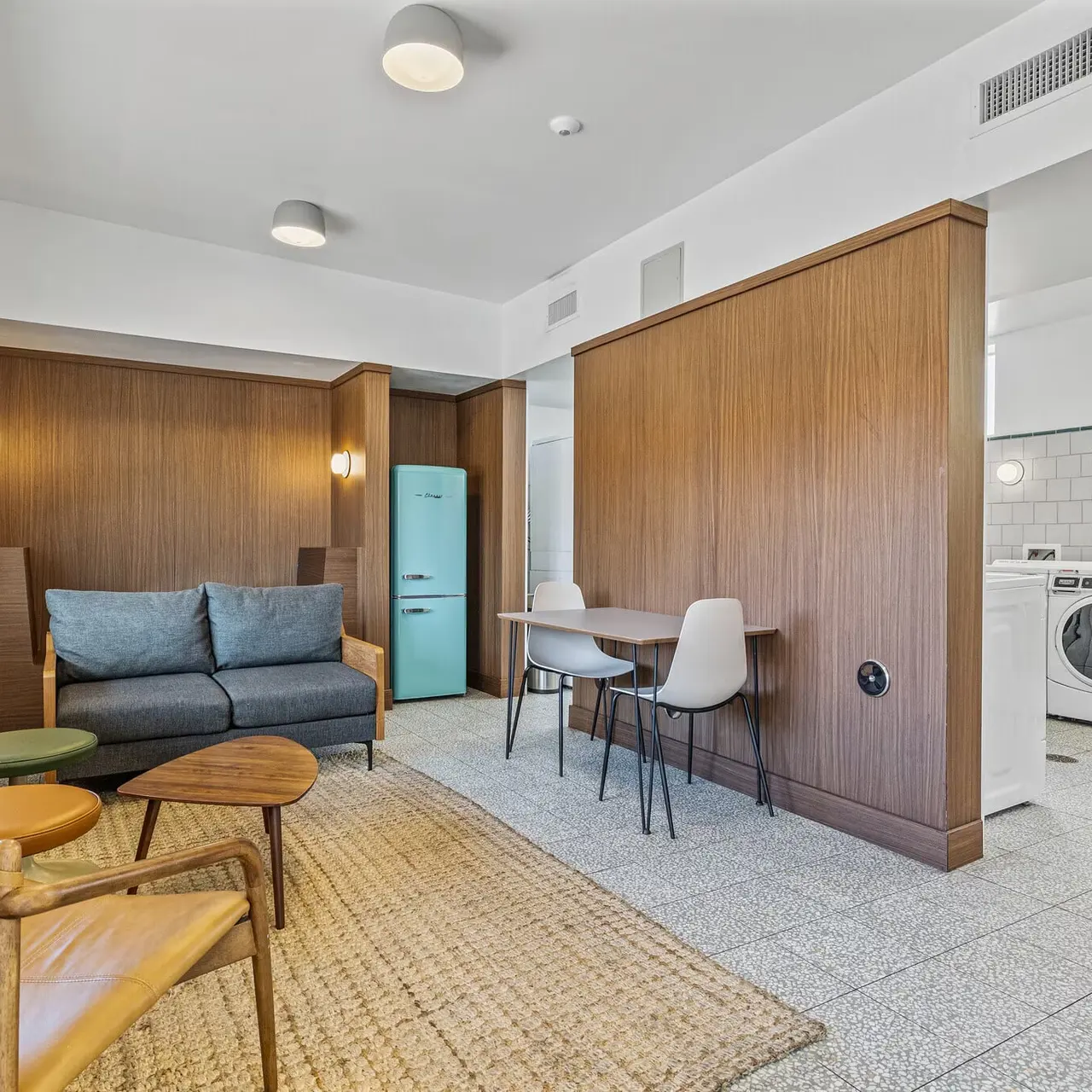 Contemporary Living Space with Laundry Area A modern living space featuring a gray sofa, wooden accents, a blue refrigerator, and a dining area with white chairs. There's a laundry area visible in the background.