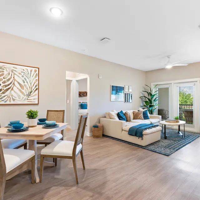 Contemporary Living Space A modern living room and dining area featuring a beige sofa, dining table with blue dishes, and a plant. The room has large windows with natural light and a decorative artwork on the wall.