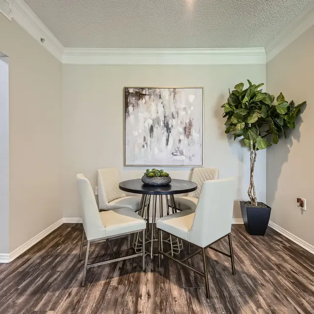 A modern dining area featuring a round black table set with four white chairs. There is a decorative plant in a pot next to the table and a wall art piece above it, all set against a light-colored wall.