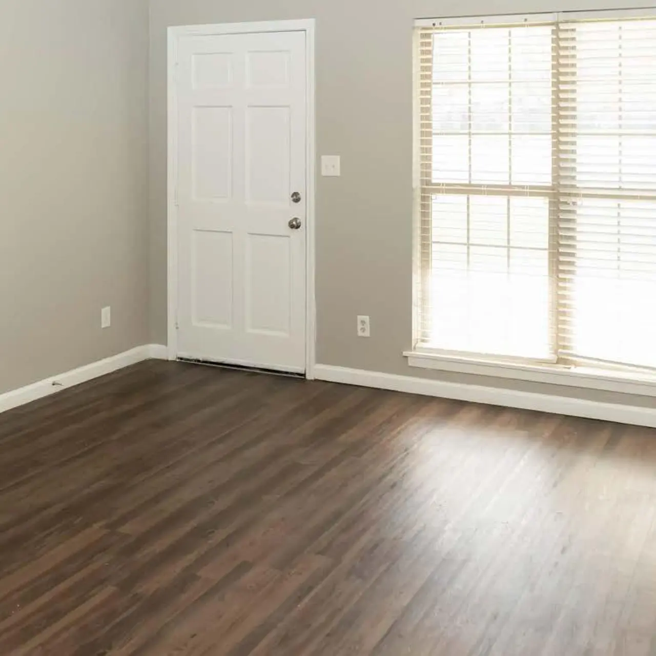 An empty room with gray walls, a door, and large windows with blinds, showcasing wooden flooring.