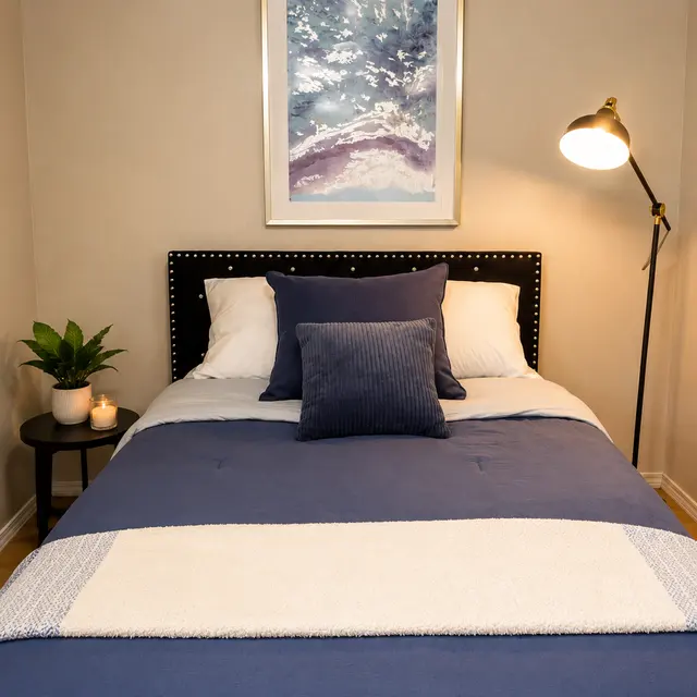 A cozy bedroom featuring a neatly made bed with navy blue bedding, decorative pillows, and a light-colored throw blanket. There is a framed artwork above the headboard and a floor lamp beside the bed, along with a small side table holding a plant and a lit candle.