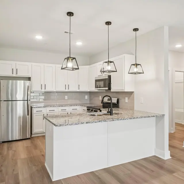 A modern kitchen featuring white cabinetry, stainless steel appliances, and a large island with a granite countertop. Pendant lights hang above the island, and the flooring is light wood.