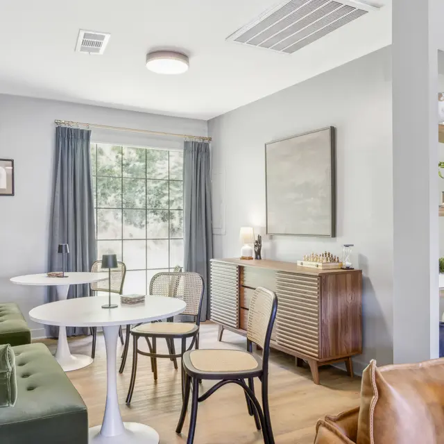 Modern living space with green sofas, round table, wooden cabinet, and large window. Bright and airy atmosphere with neutral tones.