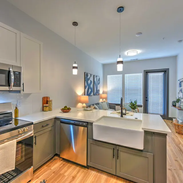 Modern Kitchen Interior A modern kitchen featuring stainless steel appliances, white cabinetry, and a farmhouse sink. The area has wooden flooring and warm lighting with an adjoining living space visible.