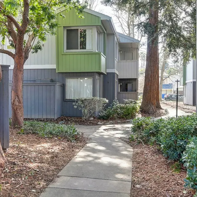 A walkway through a landscaped apartment complex featuring grey building exteriors with green accents, bordered by trees and shrubs.