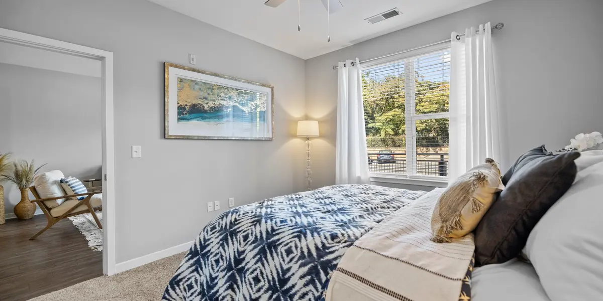 A cozy modern bedroom featuring a bed with an intricate blue and white patterned blanket. Sunlight streams in through large windows adorned with sheer white curtains. A framed artwork hangs on the wall, and a stylish floor lamp provides additional lighting. A door leads to another room, hinting at a spacious layout.