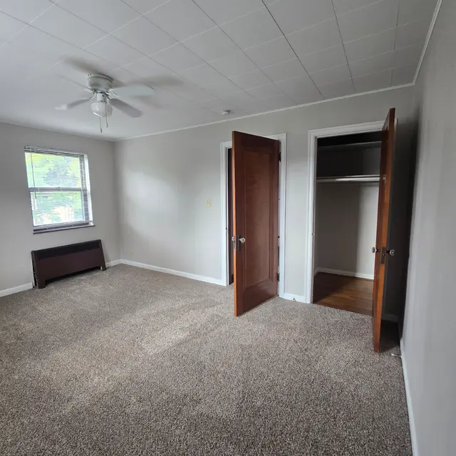 Interior View of Bedroom A spacious bedroom with beige carpet, featuring two open wooden doors leading to a closet. A ceiling fan is visible and there is a window allowing natural light into the room.