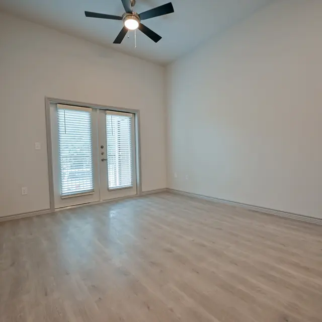 A spacious empty living room with a ceiling fan and large glass doors leading to an outdoor space. The flooring is a light wood laminate, and the walls are painted in a neutral color.