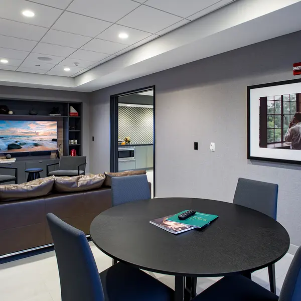 Modern lounge area with a couch, round table, and a large window; features a TV and framed art on the walls.