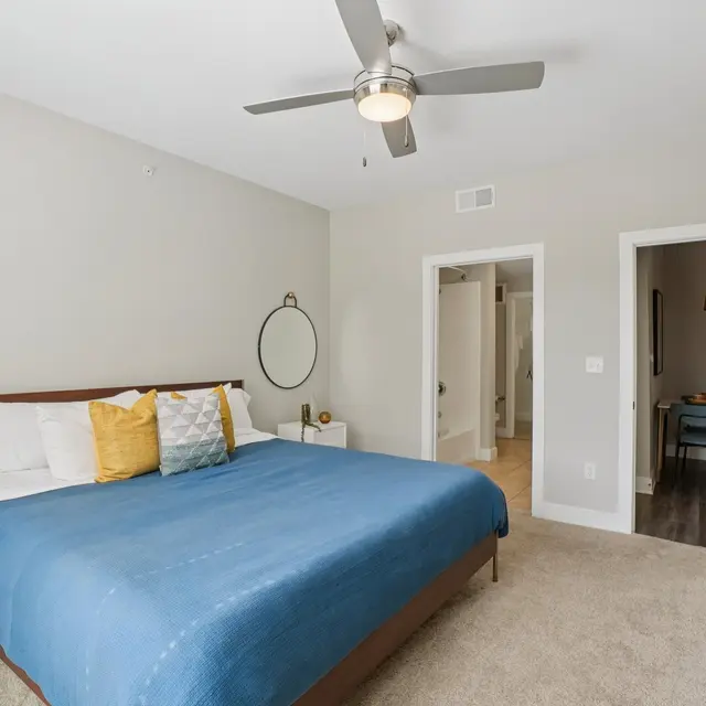 A spacious bedroom featuring a large bed with a blue blanket and decorative pillows. The room has a ceiling fan, light-colored walls, a mirror on the wall, and a doorway leading to another space. A wooden dresser is positioned next to the bed.