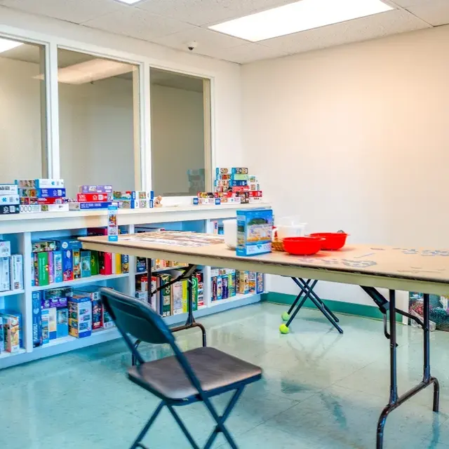 A simple room with a large table in the center, surrounded by chairs. Shelves are filled with various puzzle boxes, and there is a red container on the table.