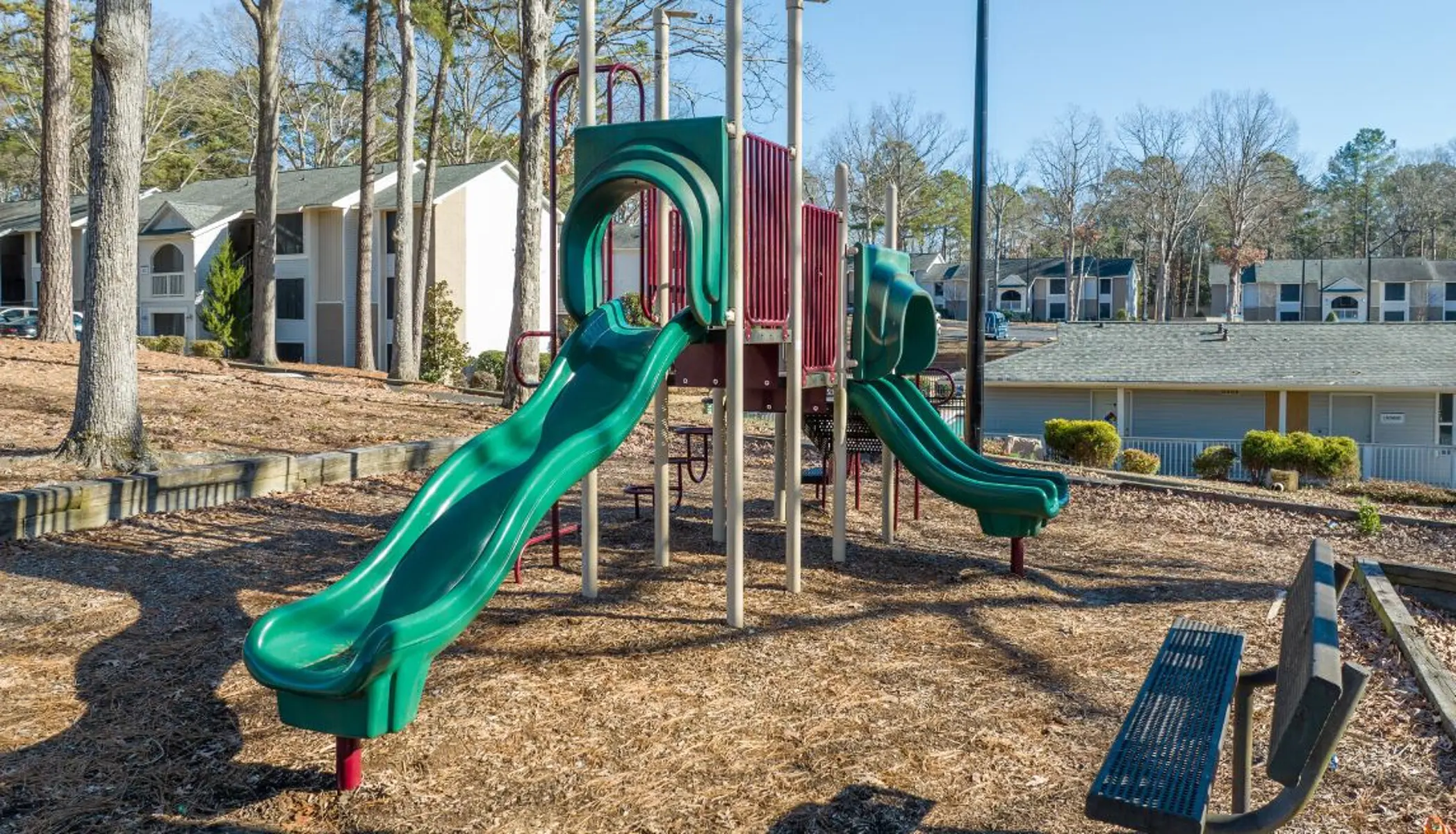 A playground featuring green slides set among trees and residential buildings in the background.