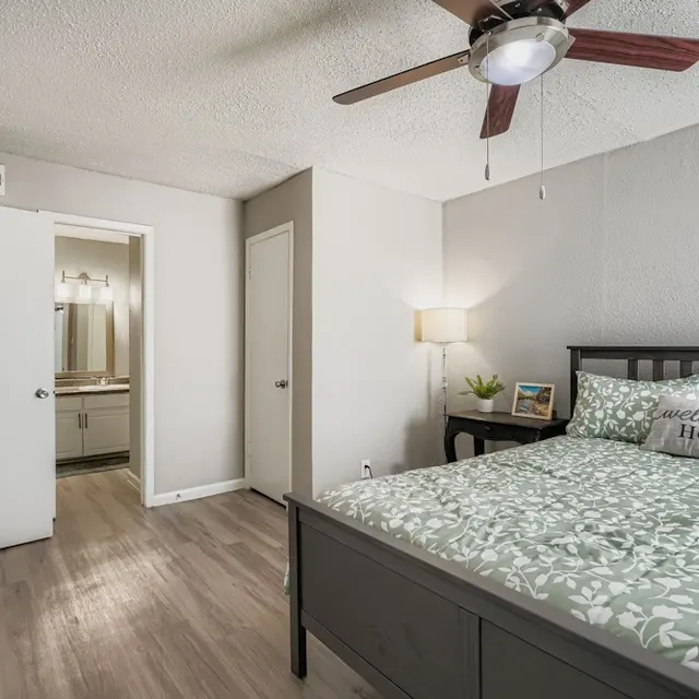 Modern Bedroom Design A modern bedroom featuring a bed with green floral bedding, a bedside table with a lamp and a framed picture, and a ceiling fan. A bathroom door is visible in the background.