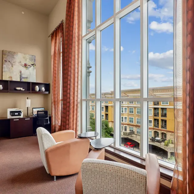 A well-lit hotel lobby area featuring large windows with a view of the outside. Inside, two chairs, a small table, and a desk setup are visible. The walls have decorative art, and rich, flowing curtains frame the windows. The aesthetic is modern and welcoming.