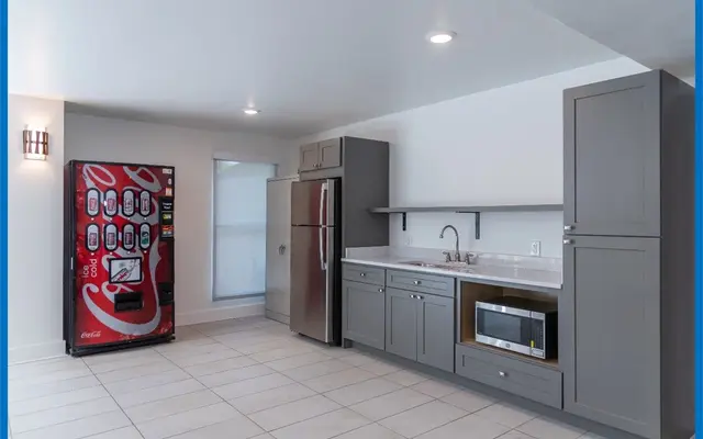 A modern kitchen space featuring a vending machine, stainless steel refrigerator, sink, microwave, and gray cabinetry against tiled flooring.