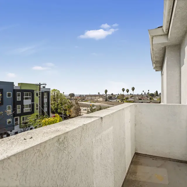 A balcony with a white railing overlooking a residential area with buildings and palm trees in the background under a clear blue sky.