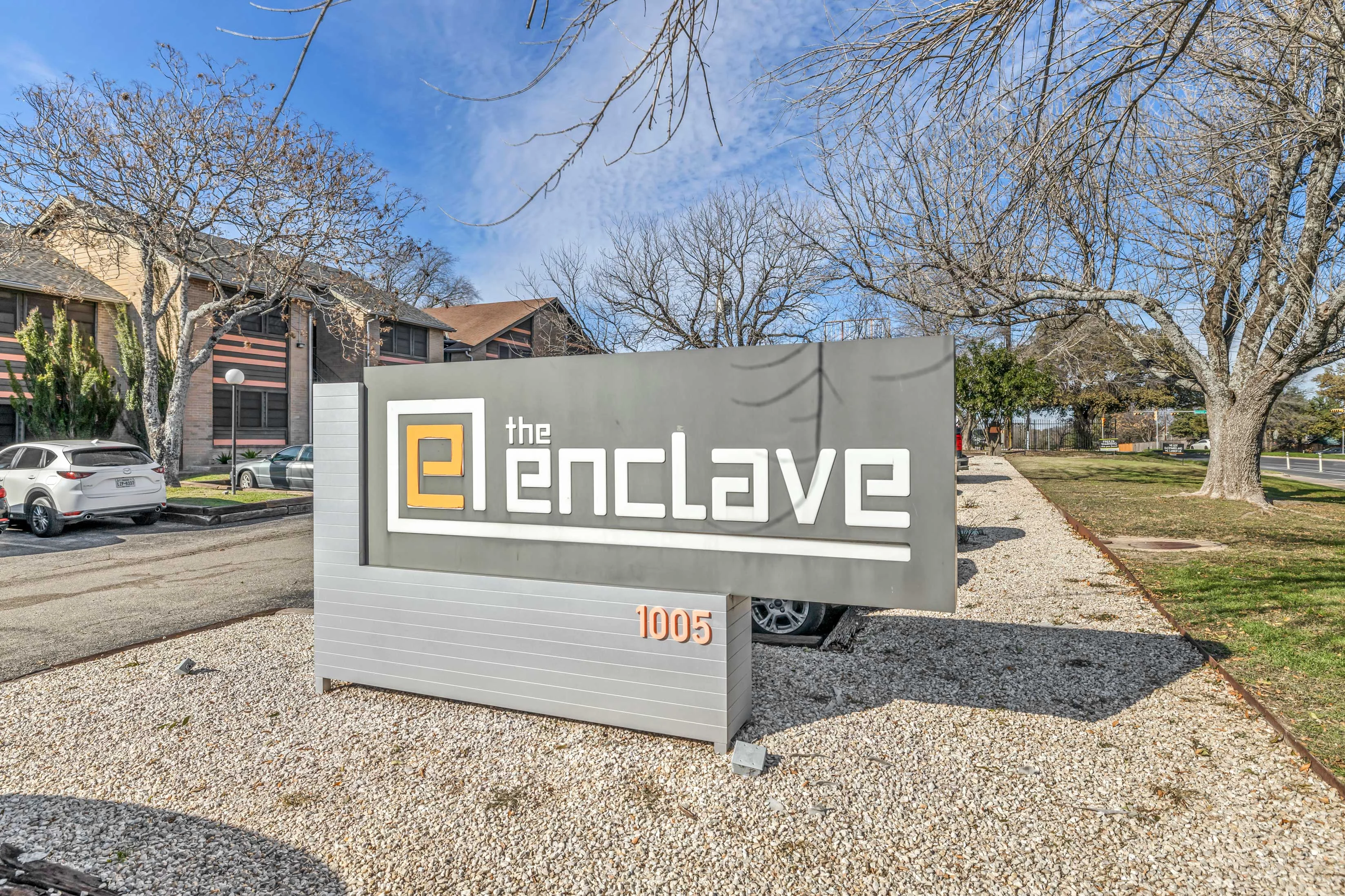 An outdoor sign of The Enclave Apartments 1005 near an apartment building in Austin, TX, resident parking, and green space with trees. 