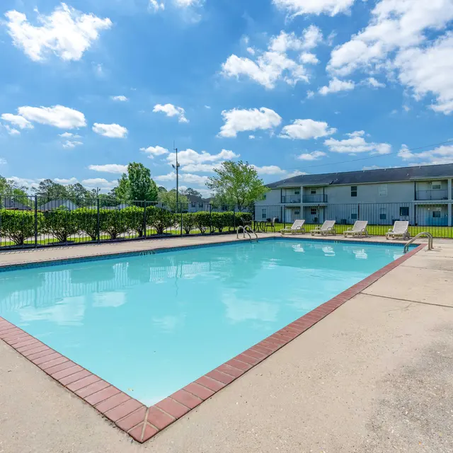 Shimmering pool surrounded by landscaped area and an apartment building in the background under a bright blue sky with scattered clouds.
