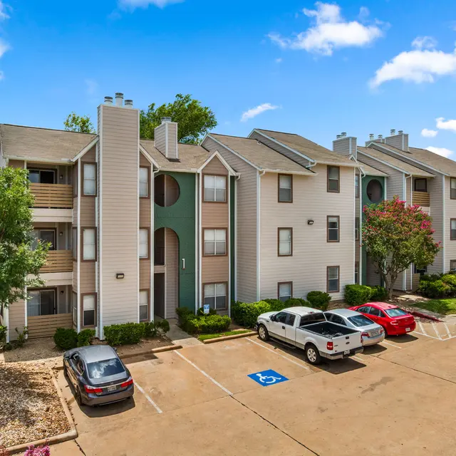 Exterior view of a multi-story apartment complex with landscaped parking area and clear blue sky.