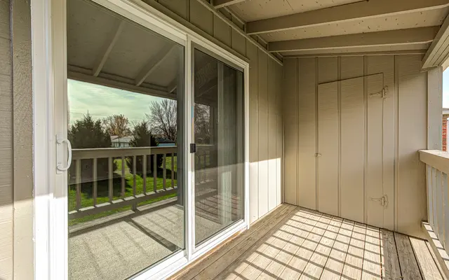 A small balcony area with sliding glass doors and wooden flooring. The balcony features a railing and is enclosed by beige walls. The reflection in the glass shows a lawn and trees outside.