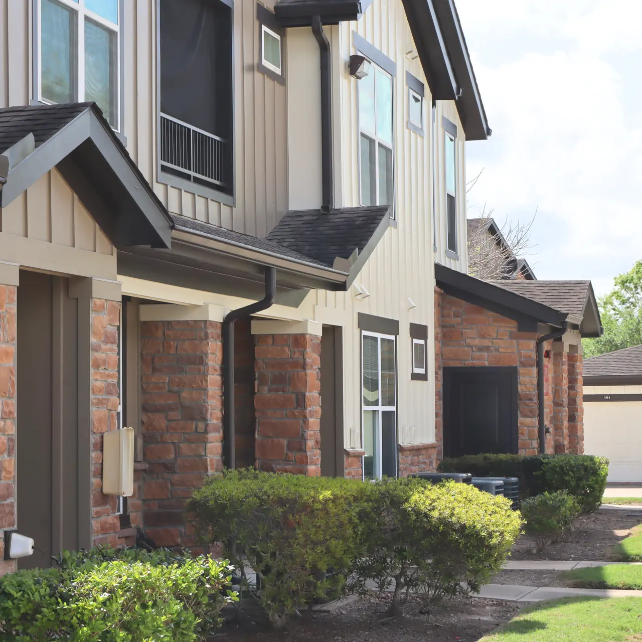 An exterior view of several apartment buildings with a landscaped path and shrubs in a sunny environment. The buildings feature a mix of stone and siding, with clear blue skies in the background.