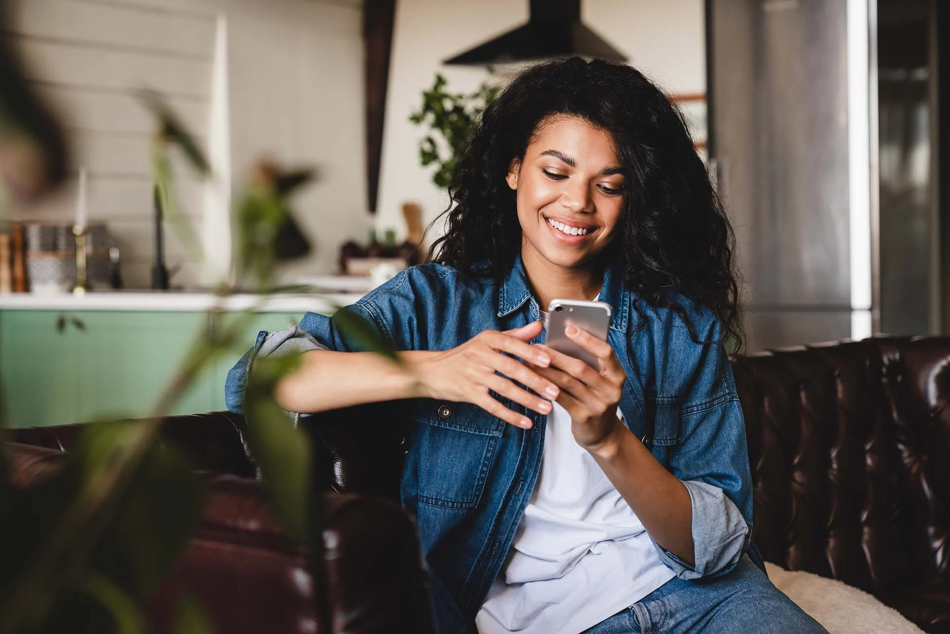 Woman enjoying her smartphone in a cozy home A smiling woman with curly hair seated on a sofa, holding a smartphone. She is dressed in a denim jacket and is looking at the phone with a cheerful expression. A cozy interior is visible in the background.