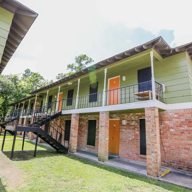Two-story apartment building with green siding and orange doors, surrounded by grass and trees.