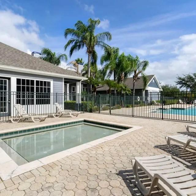 A sunny outdoor pool area featuring a hot tub, lounge chairs, and palm trees, with a swimming pool visible in the background.