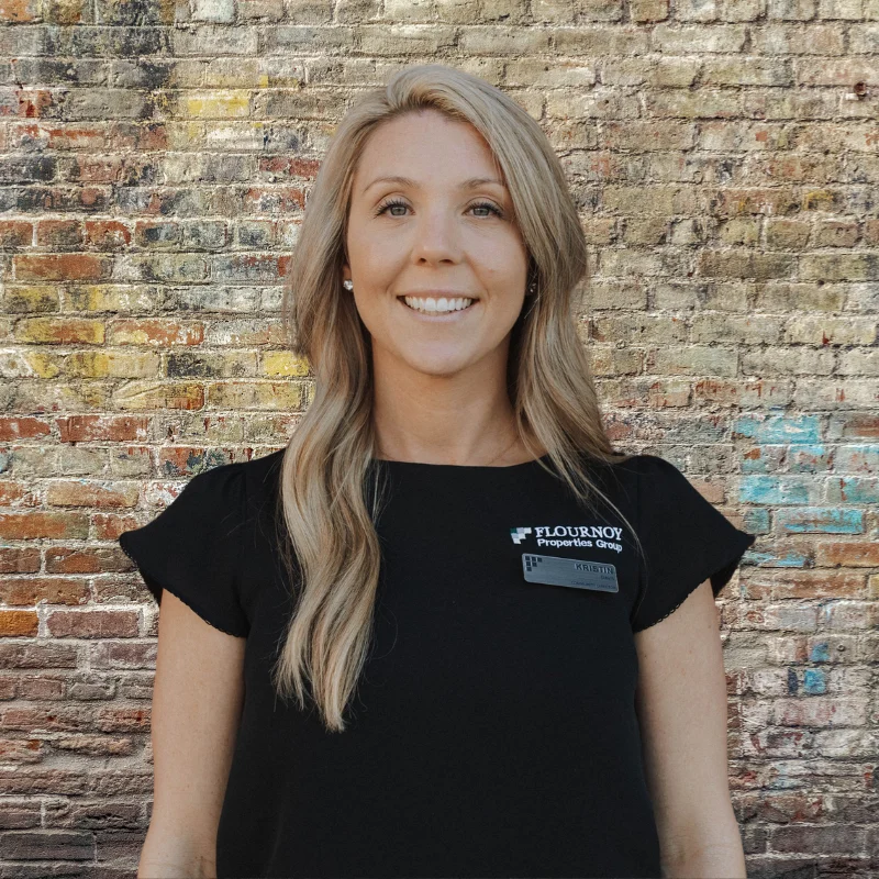 A smiling woman with long blonde hair wearing a black blouse and a name tag in front of a brick wall.