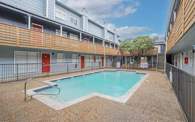 A pool area surrounded by two-story apartment buildings. The pool is rectangular with a ladder, and there are balconies above. A tree is in the background, and there are several signs near the pool.