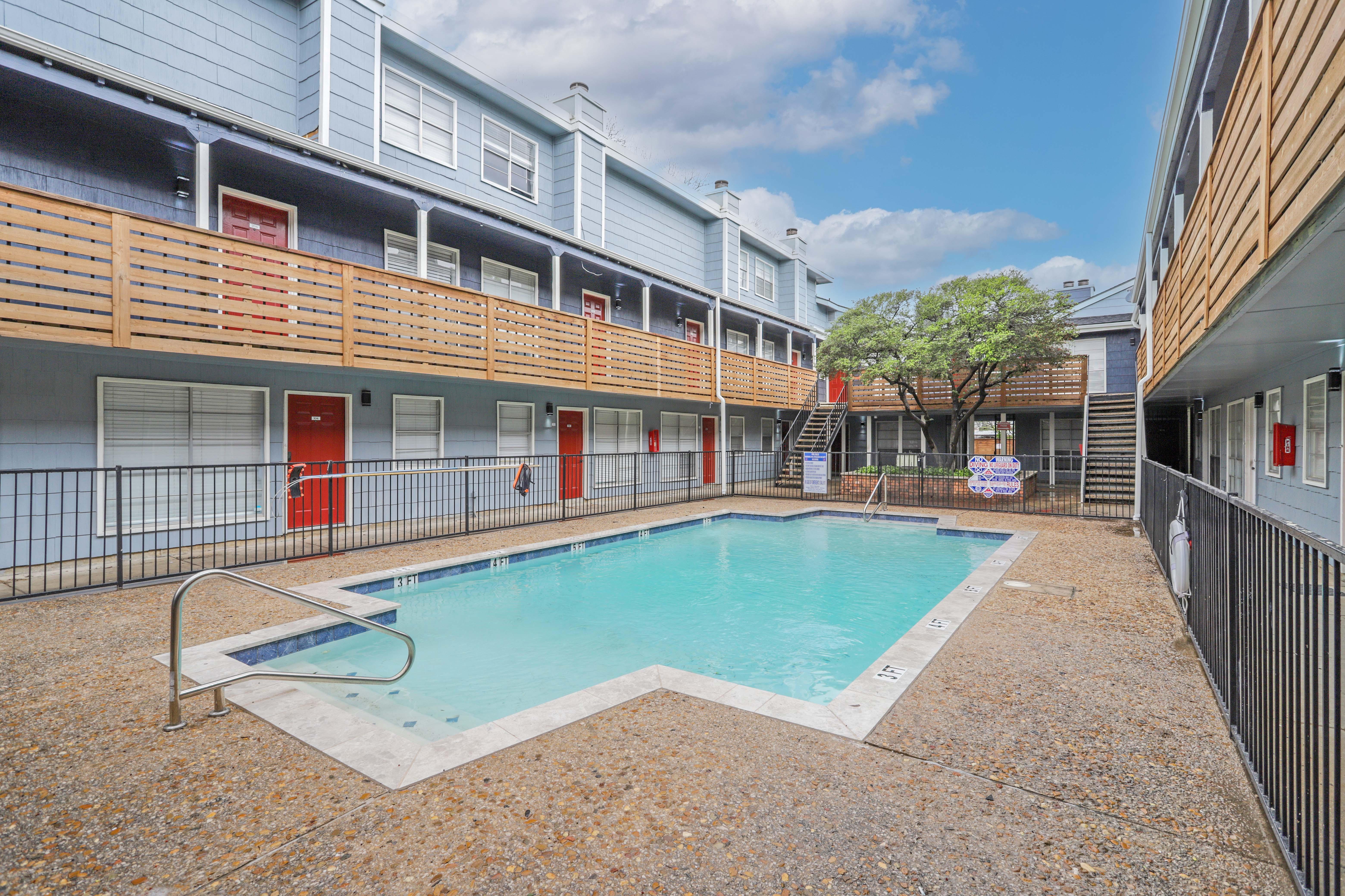 A pool area surrounded by two-story apartment buildings. The pool is rectangular with a ladder, and there are balconies above. A tree is in the background, and there are several signs near the pool.
