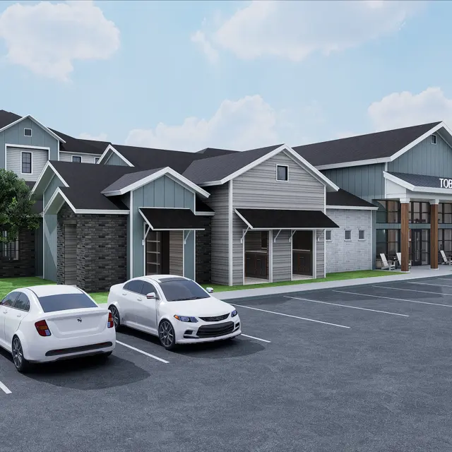 An architectural rendering of an apartment complex with modern design elements. Two cars parked in the foreground, lush green lawns, and trees visible. The building features a mix of stone and siding materials with a characteristically sloped roof.