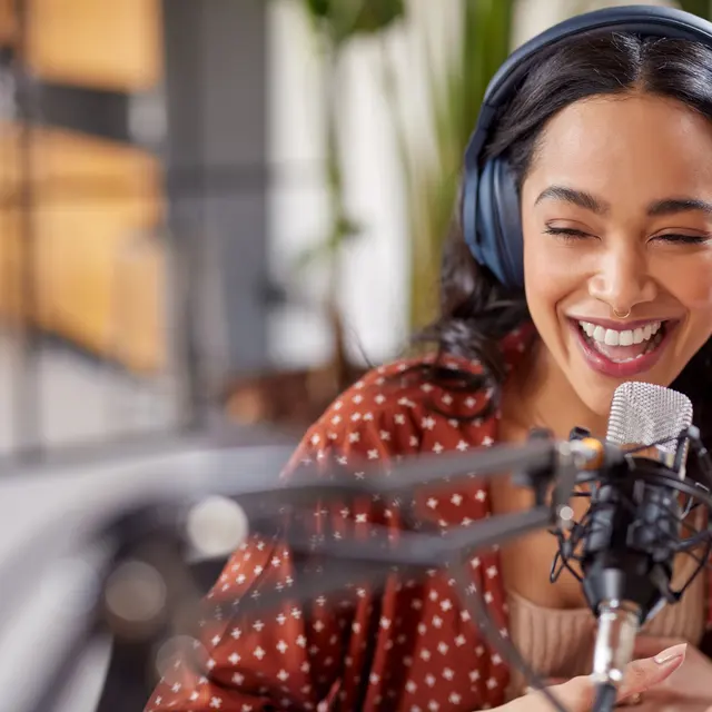 A woman with long dark hair, wearing headphones, laughing while speaking into a microphone in a modern studio setting.