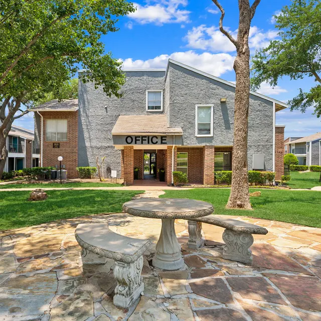 An office building of an apartment complex surrounded by green grass and trees, featuring a stone bench area in front.