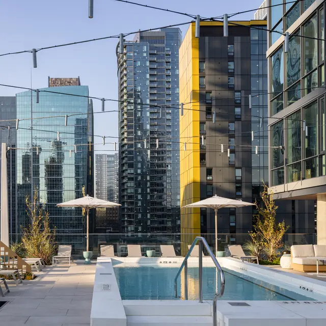 A modern rooftop pool area surrounded by high-rise buildings, featuring lounge chairs, umbrellas, and greenery.