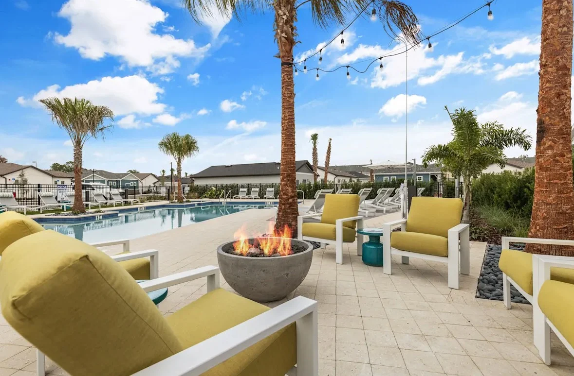 A scenic poolside area featuring a fire pit surrounded by modern yellow armchairs, palm trees, and string lights in the background.