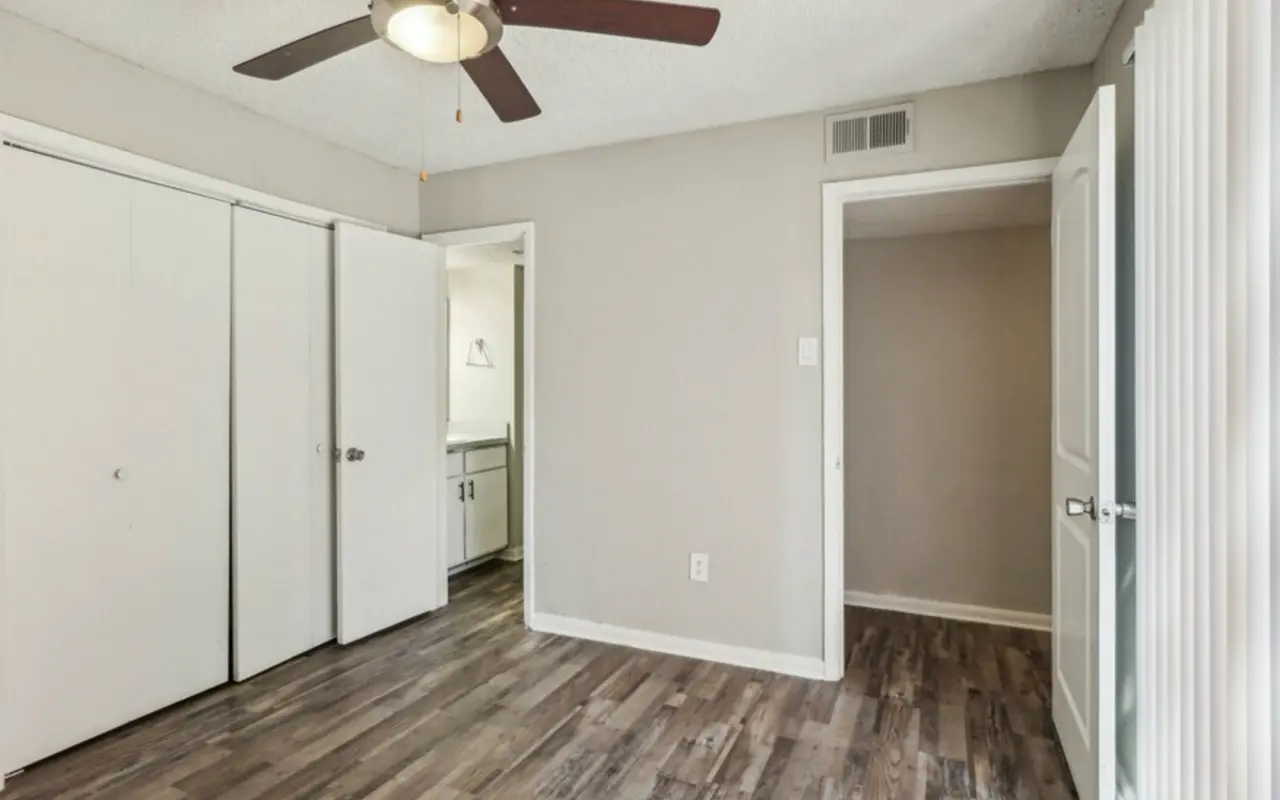 A spacious bedroom featuring light brown wooden floors, a ceiling fan, and gray painted walls. There are two white sliding door closets on one side and an open door leading to another room. Natural light is visible from a window on the left.