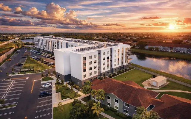 Aerial view of a modern apartment complex by a lake at sunset, with solar panels on the roof and parked cars.