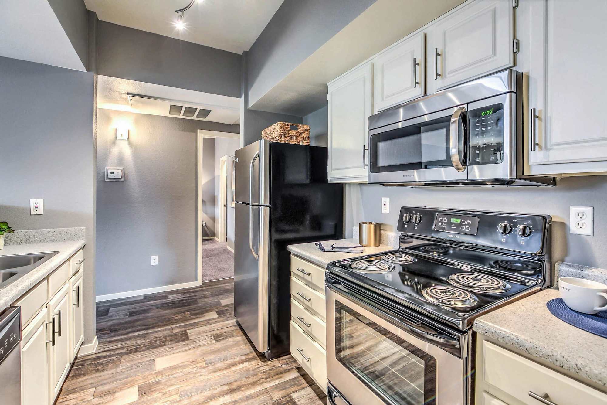 A modern kitchen featuring stainless steel appliances, including a microwave and oven. The decor includes light-colored cabinetry and gray walls, with a view into an adjoining room.