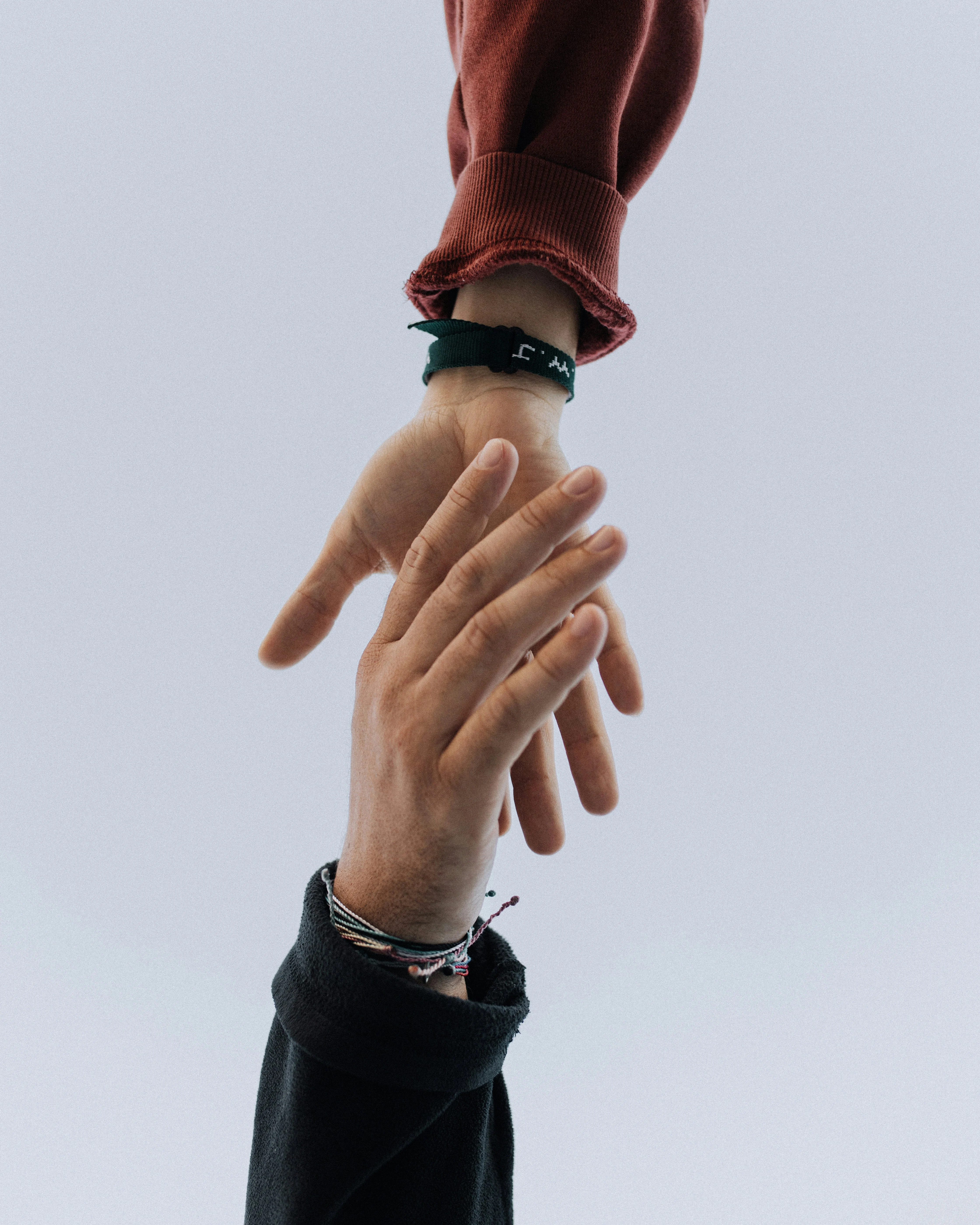 Symbolic Connection: Reaching Hands Two hands reaching towards each other against a light background. The top hand wears a red long-sleeve shirt and a green wristband, while the bottom hand has several bracelets and is wearing a dark outfit.