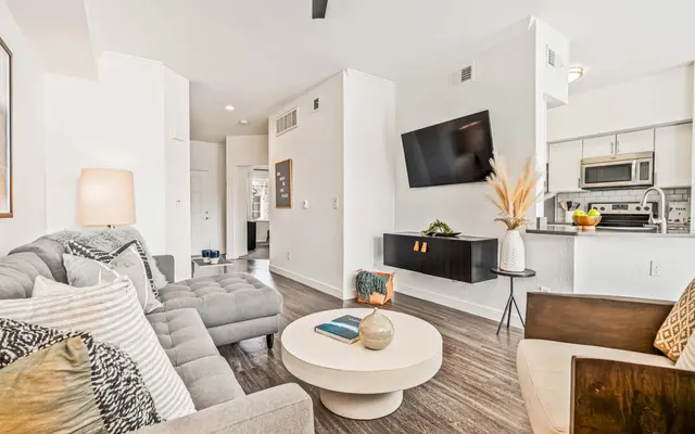 A modern living room featuring a gray sectional sofa, a round coffee table, and a mounted television. The space is well-lit with a floor lamp and offers a view of the kitchen area.