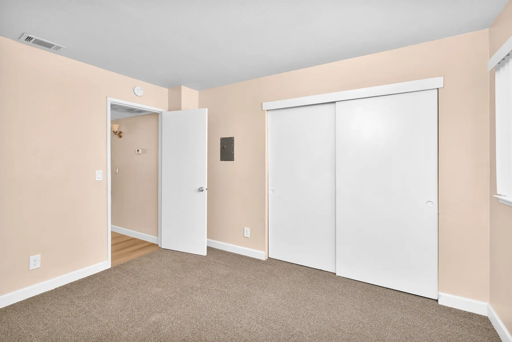 An empty room with beige walls and carpeted floor. There is a closet with sliding white doors on one wall and a partially open door leading to another room. Natural light comes in from a window.
